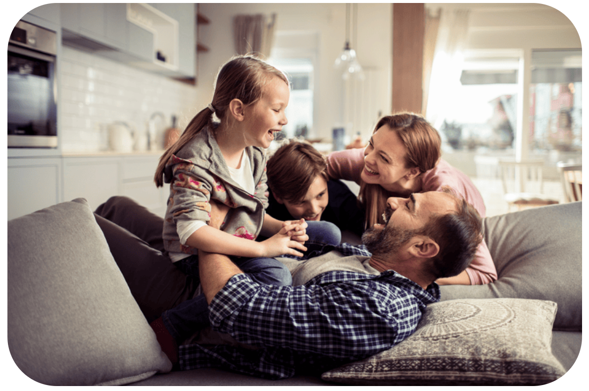 Father lying on a couch as his kids and partner pile on him, all smiling and laughing in a cozy living room.