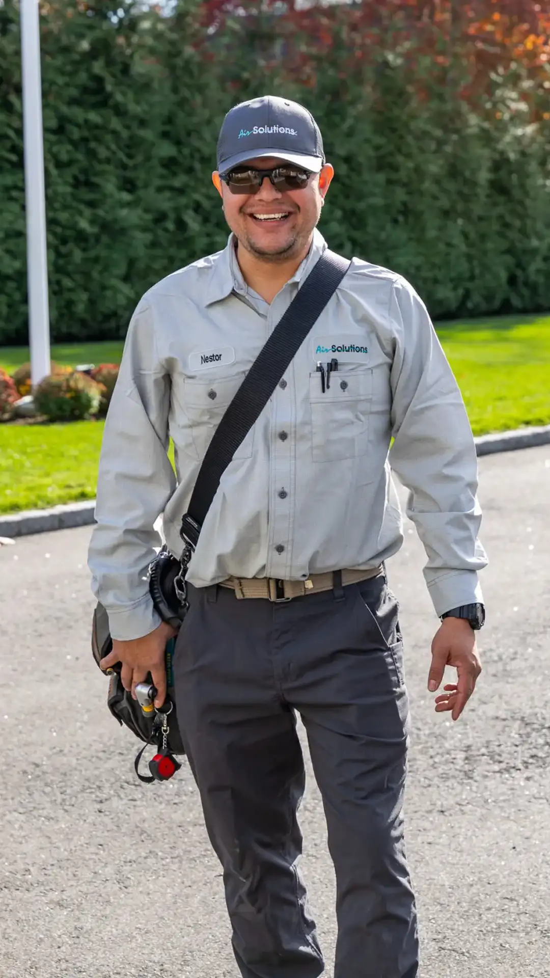 Smiling technician in a gray uniform with name tag “Nestor,” wearing a cap and crossbody bag outdoors with tools visible.